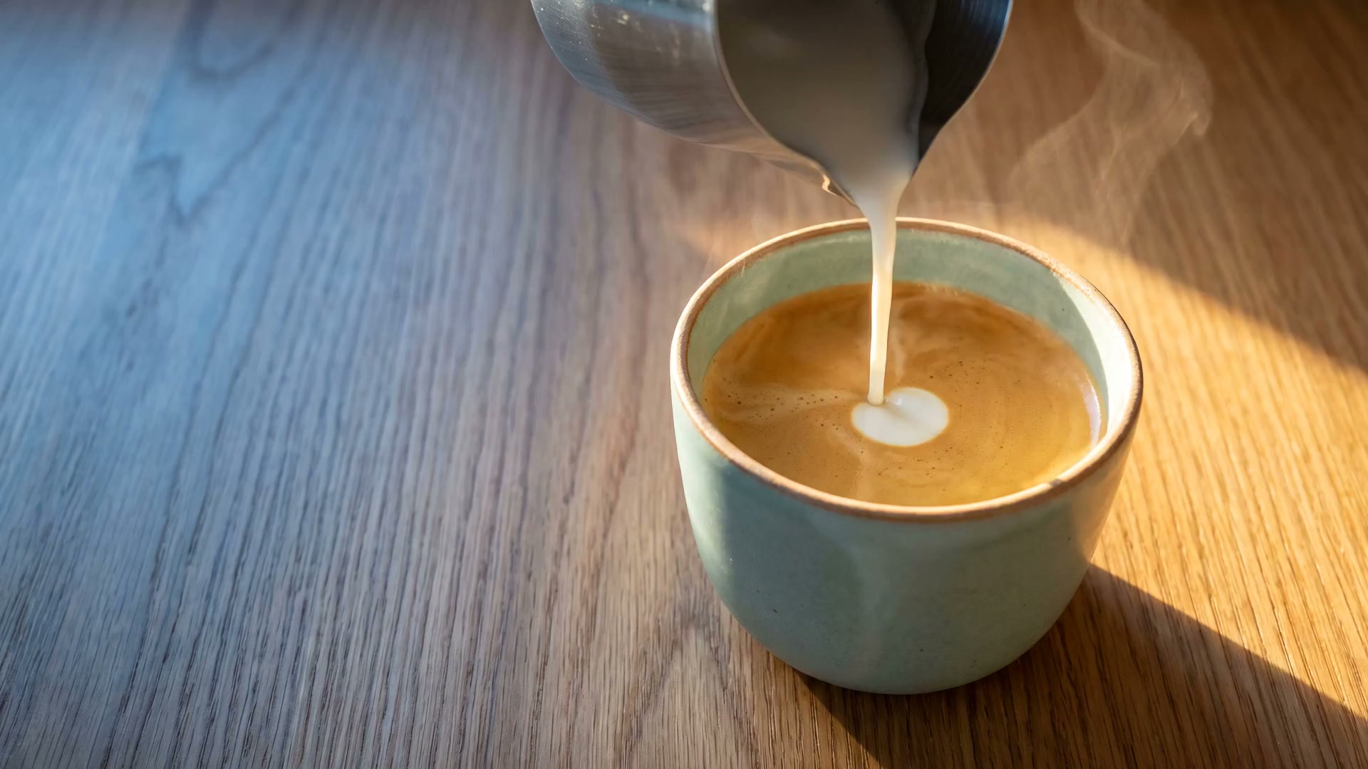 Steamed milk pouring into an espresso in a green-blue stoneware cup, in early morning light